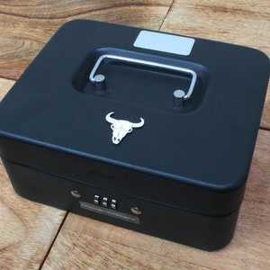 Puede incluir: Caja de metal negra con una cerradura de combinación y un diseño de calavera en la tapa. La caja tiene una manija de transporte.