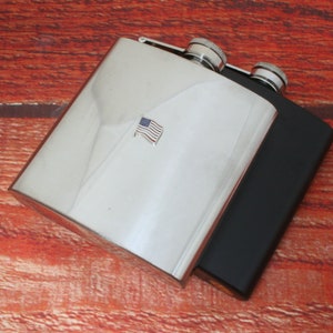 May include: Two stainless steel hip flasks, one with a small American flag design. The flasks are on a wooden surface.