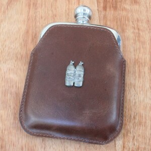 May include: A brown leather-covered hip flask with a silver metal top and a silver metal emblem of two scuba diving tanks on the front.