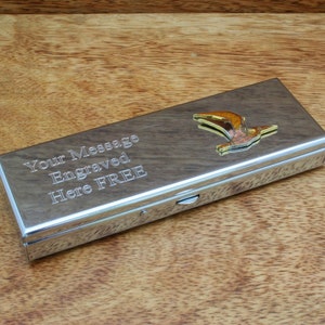 Puede incluir: Una caja de metal plateado con una tapa con bisagras. La caja tiene un diseño de pájaro dorado en la parte superior y el texto "Your Message Engraved Here FREE" está grabado en la parte delantera.