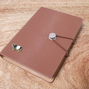 Puede incluir: Un diario de cuero marrón con un pin de vaca blanco y negro y un cierre de botón plateado. El diario tiene una superficie texturizada y está atado con una cuerda marrón.