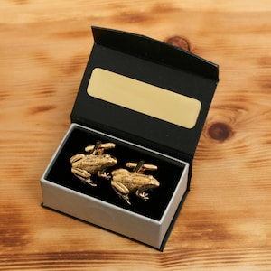 Peut inclure: Une paire de boutons de manchette dorés en forme de grenouille, présentée dans un coffret noir avec une plaque dorée. Les boutons de manchette ont une peau texturée et sont sur un intérieur en velours noir.