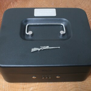 May include: Black metal cash box with a combination lock and a silver handle. The top of the box has a silver rifle emblem and the words "Safe Hunter Equipment" engraved on a silver plate.