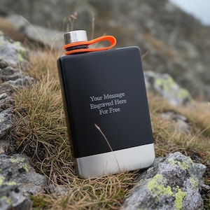 Peut inclure: Une flasque noire avec une base et un bouchon argentés, et une boucle orange. La flasque porte l'inscription "Your Message Engraved Here For Free". La flasque est rectangulaire et semble être en métal.