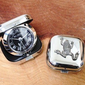 May include: A silver-coloured travel clock with a black face and Roman numeral markings. The clock is open and the lid is closed. The lid has a silver-coloured frog design.