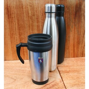 Peut inclure: Une tasse à café isotherme argentée avec une poignée et un couvercle noirs. La tasse a un petit emblème argenté avec un motif rouge et bleu. La tasse est à côté de deux bouteilles d'eau en acier inoxydable, une argentée et une noire.