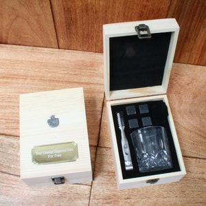 May include: A wooden box with a black velvet interior contains a set of whiskey stones, a pair of tongs, and a glass. The box has a brass latch and a gold plaque that says "Your message engraved here for free".
