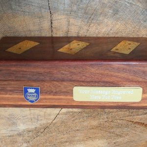 Puede incluir: Una caja de madera con una placa dorada que dice "Su mensaje grabado aquí gratis". La caja tiene un escudo azul y dorado con las letras "MN" en su interior. La parte superior de la caja tiene tres dominós incrustados.