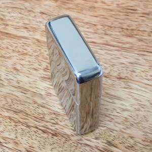 May include: A silver lighter with a smooth, polished finish. The lighter is closed and sitting on a wooden surface.