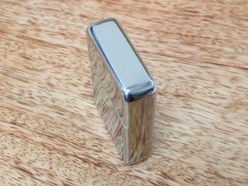 May include: A silver lighter with a smooth, polished finish. The lighter is closed and sitting on a wooden surface.