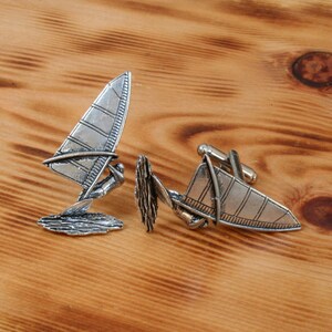 May include: A pair of silver cufflinks featuring a windsurfer design. The cufflinks are detailed with a windsurfer and waves.