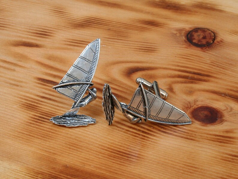 May include: A pair of silver cufflinks featuring a windsurfer design. The cufflinks are detailed with a windsurfer and waves.