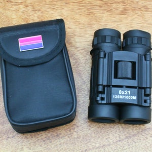 May include: Black binoculars with a carrying case. The binoculars have a pink, purple, and blue stripe on the front. The binoculars are labeled "8x21 126M/1000M".