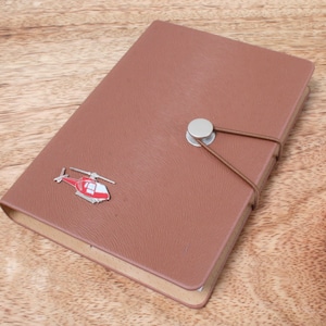 Könnte beinhalten: Ein braunes, ledergebundenes Notizbuch mit einem silbernen Knopfverschluss und einem kleinen roten Hubschrauberdesign auf dem Einband.