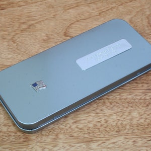May include: A rectangular, silver-colored metal tin with a small American flag emblem on the top. The tin has a label with text on it. The tin is on a wooden surface.