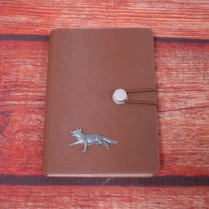 May include: A brown leather journal with a silver fox design and a silver clasp. The journal is on a red wooden background.