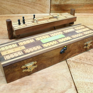 May include: A wooden cribbage board with a hinged lid and brass clasps. The board has a light brown wood finish with a darker brown wood inlay. The board is open and shows the playing surface with a green label that says "Cribbage".
