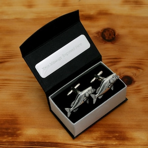 Può includere: Un paio di gemelli a forma di pesce argentati presentati in un cofanetto nero con interno bianco. Il coperchio della scatola è aperto, rivelando i gemelli e un'etichetta argentata con la scritta "Your Message Engraved Here".