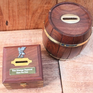 Pode incluir: Duas caixas de madeira para guardar dinheiro. Uma é uma caixa quadrada com uma figurinha de fada na tampa e uma fenda de latão para moedas. A outra é uma caixa com forma de barril com uma fenda de latão para moedas e bandas de latão ao redor do meio. O texto na caixa quadrada diz "Sua mensagem gravada aqui de graça".