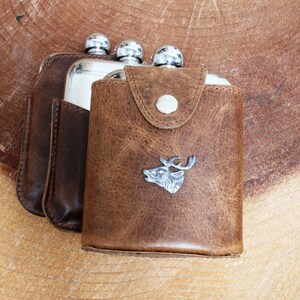 May include: Two stainless steel hip flasks in a brown leather case with a deer head design.