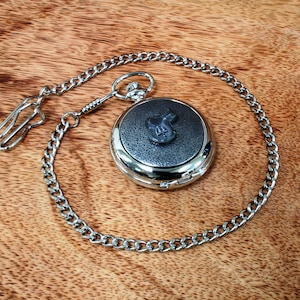 May include: A silver pocket watch with a black leather face and a silver chain. The watch face has a small silver design in the center.