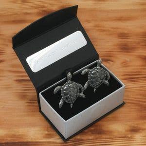 May include: A black gift box with a silver inscription that reads "Your text or design here". The box is open and contains two silver turtle cufflinks.