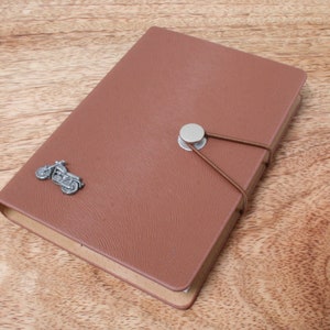 May include: A brown leather-bound journal with a silver motorcycle charm and a silver button closure. The journal is closed and lying on a wooden surface.