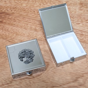 Puede incluir: Una caja de pastillas de plata con un diseño de árbol de la vida en la tapa. La caja está abierta y tiene dos compartimentos en el interior.