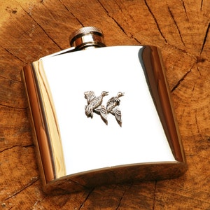 May include: A silver flask featuring a design of two ducks in flight. The flask is resting on a wooden surface.