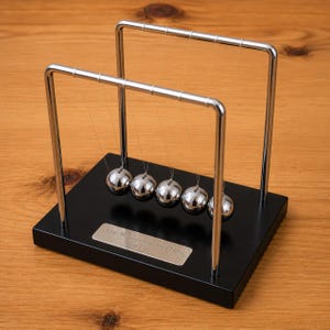 Può includere: Un pendolo di Newton con cinque sfere metalliche argentate lucide sospese tramite fili sottili a una cornice cromata. Il dispositivo è montato su una base rettangolare nera con una targhetta color argento incisa con il testo Your Message Engraved Here For Free.