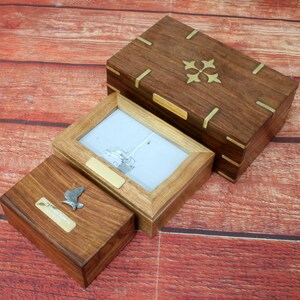 May include: Three wooden boxes with brass accents. The largest box has a decorative brass design on the lid. The middle box has a clear glass insert and a brass plaque. The smallest box has a brass bird figurine on the lid.