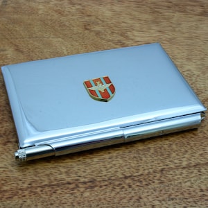 Op de afbeelding: Een zilveren metalen kaarthouder met een rood, wit en blauw embleem met een gevleugelde adelaar. Het embleem is bevestigd aan de voorkant van de kaarthouder.