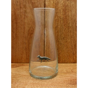 Può includere: Una caraffa di vetro trasparente con una statuetta di volpe in peltro all'interno. La caraffa ha una base larga e un collo stretto.