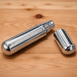 Puede incluir: Una petaca plateada pulida con forma rectangular redondeada y tapa extraíble. La petaca tiene el texto grabado "Your Message Here" y está sobre una superficie de madera. La tapa está al lado de la petaca.