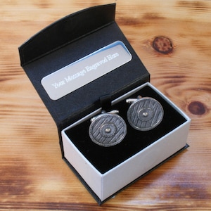 May include: A pair of silver-toned cufflinks with a shield design, presented in a black gift box with a white interior. The box has a silver plate that reads "Your Message Engraved Here".