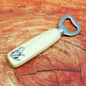 May include: A wooden bottle opener with a silver frog design on the handle. The opener has a silver metal loop for opening bottles.