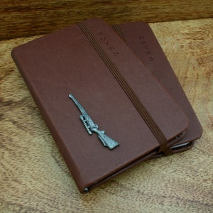 Puede incluir: Dos cuadernos de cuero marrón con la palabra "NOTES" grabada en la portada. Un dije de rifle plateado está colocado en la parte delantera de un cuaderno. Los cuadernos están encuadernados con una banda elástica marrón.