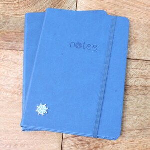 May include: Two blue notebooks with a white elastic band closure. The top notebook has the word "notes" printed on the front cover. A small silver compass is attached to the front cover of the bottom notebook.