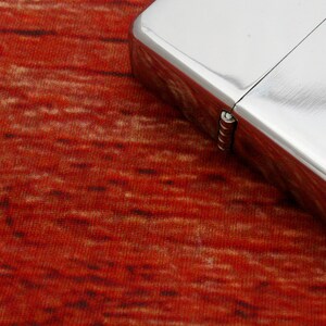 Puede incluir: Un encendedor plateado con una bisagra sobre una superficie texturizada roja y marr&oacute;n.