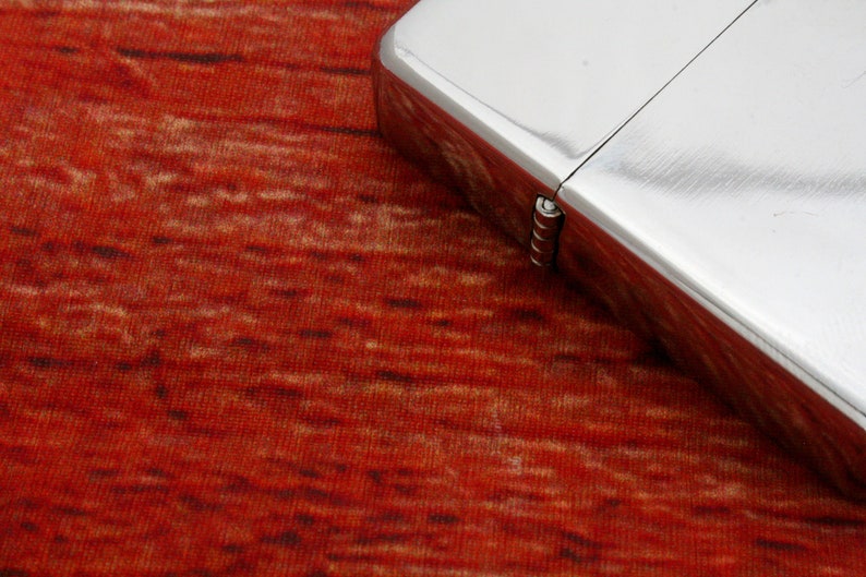 Puede incluir: Un encendedor plateado con una bisagra sobre una superficie texturizada roja y marr&oacute;n.