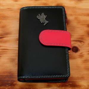 May include: A black leather wallet with a pink flap and a silver metal emblem. The wallet has a stitched design around the edges.