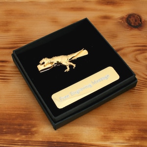 May include: A gold-colored T-Rex tie clip presented in a black box. The tie clip features a detailed dinosaur design. A gold-colored rectangular plate with the text "Free Engraving Message" is also in the box.
