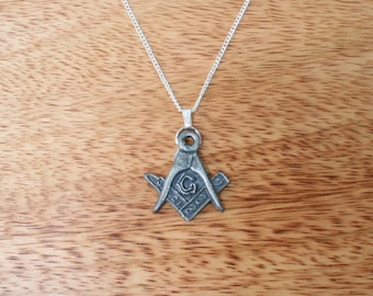 May include: A silver-toned pendant necklace featuring a Masonic symbol. The pendant depicts a square and compass with a letter "G" in the center, suspended from a delicate silver chain. The necklace is displayed against a wooden background.
