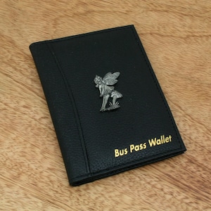 Peut inclure: Porte-carte de bus en cuir noir avec un emblème de fée argenté. Les mots "Bus Pass Wallet" sont imprimés en lettres dorées. Le portefeuille est fermé et posé sur une surface en bois.