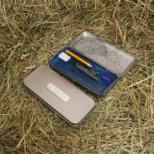 以下が含まれることがあります： コンパス、鉛筆、消しゴム、鉛筆削りが入った銀色の金属製のペンケース。ケースには「Your Name Here」というテキストが書かれた銀色のラベルが付いています。