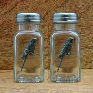May include: A set of two glass salt and pepper shakers with silver lids. Each shaker features a pewter budgie figurine inside.
