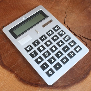 May include: A silver calculator with black buttons and a black display. The calculator has basic functions such as addition, subtraction, multiplication, division, percentage, square root, and memory functions.