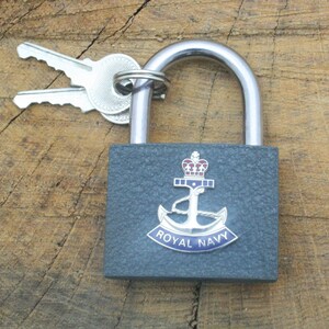 Puede incluir: Un candado gris con un grillete plateado y dos llaves. El candado tiene un emblema de la Royal Navy con una corona, un ancla y las palabras "Royal Navy".