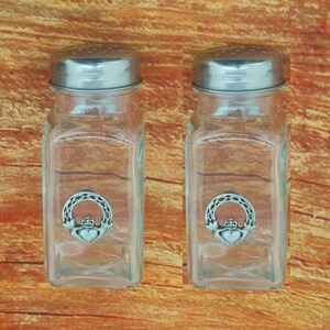May include: Two clear glass salt and pepper shakers with silver-colored metal lids. Each shaker has a decorative silver-colored emblem of a heart, crown, and hands. The shakers are rectangular and sit on a wooden surface.