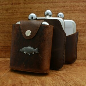 Könnte beinhalten: Drei Edelstahl-Schnapsflaschen mit braunen Ledertaschen. Die Flasche links hat ein silbernes Fischdesign auf der Tasche.